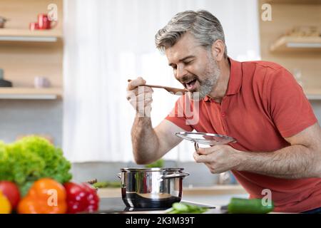 Schöner Mann mittleren Alters, der Essen schmeckte, Kücheneinrichtung Stockfoto