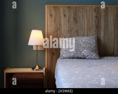 Ein Bett mit grauen Bezügen und einem hölzernen Kopfteil neben einem Nachttisch mit einer beleuchteten Lampe Stockfoto