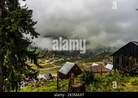Von oben Häuser und Nadelbäume am grünen Hang des Berges gegen blauen Himmel mit dunkelgrauen Wolken und Nebel am stürmischen Tag im Hochland gelegen Stockfoto