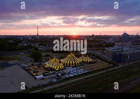 Luftaufnahme des Stadtbildes mit modernen Gebäuden und Zirkuszelten, die bei Sonnenuntergang in Deutschland in der Nähe der Straße aufgestellt wurden Stockfoto