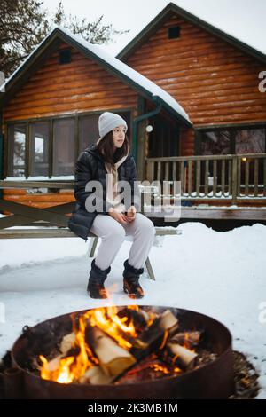 Ganzkörperfrau in Oberbekleidung schaut weg, während sie am Wintertag auf der Bank in der Nähe von Brazier mit brennenden Holzstämmen vor dem Holzhaus auf dem Land sitzt Stockfoto