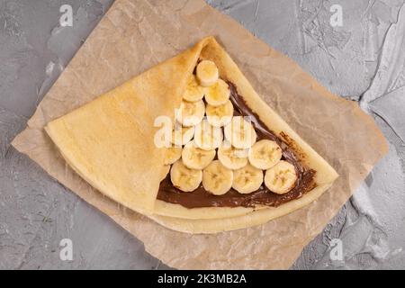 Crepes gefüllt mit Schokoladenaufstrich und Banane auf weißem Teller. Dünne Pfannkuchen, Blini. Süßes Dessert. französische Küche von oben Stockfoto