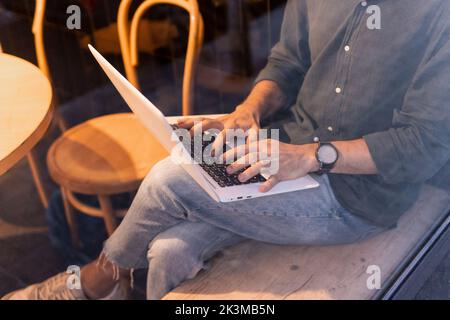 Beschnittenes, nicht erkennbarer Mann im schwarzen Hemd, der Daten auf Netbook durchstöberte, während er hinter dem Café-Fenster saß und an einem freiberuflichen Projekt arbeitete Stockfoto