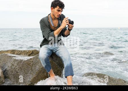 Junger hispanischer Mann in legerer Kleidung sitzt auf Stein und fotografiert am bewölkten Tag das winkende Meer Stockfoto
