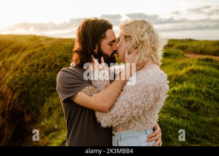 Bärtiger Mann umarmt und küsst die blonde Freundin, während er bei Sonnenuntergang in Aviles, Spanien, auf einer grasbewachsenen Klippe steht Stockfoto