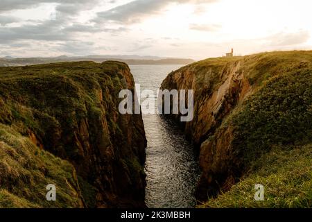 Atemberaubender Blick auf die Bucht mit plätscherndem Wasser und Klippen mit Leuchtturm, der sich gegen den bewölkten Sonnenuntergang in Aviles, Spanien, befindet Stockfoto