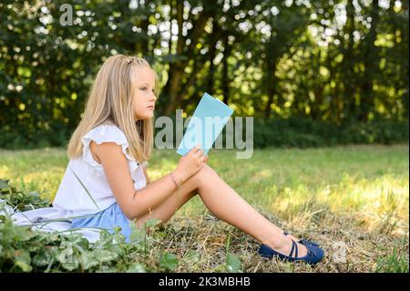 Ganzkörper-Seitenansicht blondes Mädchen in lässiger Kleidung sitzt weg auf Decke in der Nähe von Bäumen und lesen dünne Buch, während verbringen Sommerwochenende Tag in Stockfoto