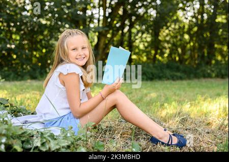 Ganzkörper-Seitenansicht blondes Mädchen in legerer Kleidung sitzend Blick auf Kamera auf Decke in der Nähe von Bäumen und lesen dünne Buch während des Sommers Wochenende d Stockfoto