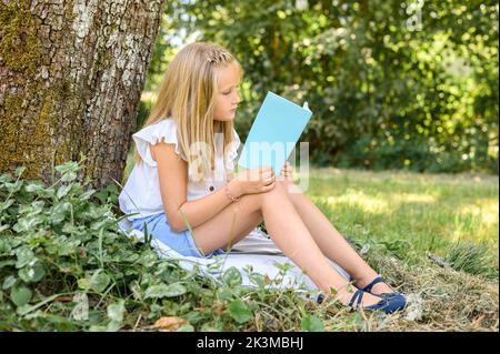 Ganzkörper-Seitenansicht blondes Mädchen in legerer Kleidung, das auf einer Decke in der Nähe von Baum sitzt und dünnes Buch liest, während es den Sommerwochenende im Park verbringt Stockfoto
