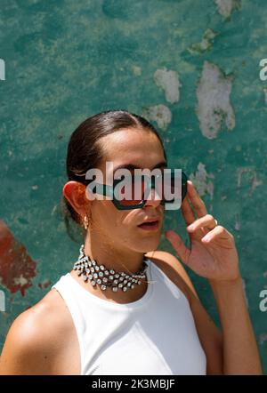 Selbstbewusstes junges, ethnisches Frauenmodell mit dunklem Haar, das modische Sonnenbrillen berührt und an sonnigen Tagen gegen eine schäbige Wand blickt Stockfoto
