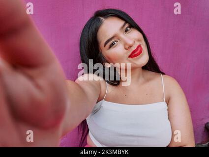 Fröhliche dicke junge Frau in stilvollem weißen Top mit langen schwarzen Haaren und roten Lippen lächelnd und Blick auf die Kamera, während Selfie gegen rosa Wand Stockfoto