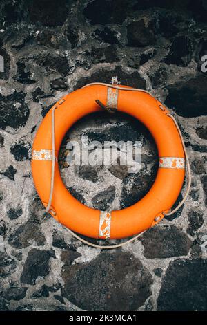 Orangefarbene Rettungsboje, die an einem Pflock an einer rauen Steinwand eines alten Gebäudes am Strand hängt Stockfoto
