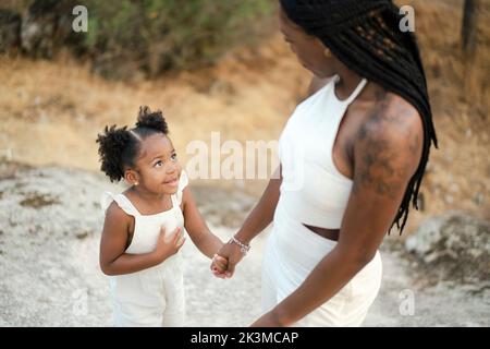 Nette fröhliche kleine afroamerikanische Mädchen in stilvollen weißen Kleid lächelnd und halten Hand der Ernte schwarze Mutter mit langen dunklen Zöpfen im Park stehen Stockfoto