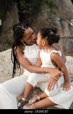Glückliche schwarze Mutter mit Afro-Zöpfen lächelt und spricht mit fröhlichen Tochter in weißen Kleidern, während sie den Sommerwochenende auf dem Land verbringt Stockfoto