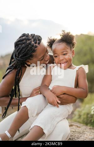 Fröhliche afroamerikanische Mutter mit lockigen Haaren umarmt und kitzelt glückliche Tochter, während Sommer Wochenende Tag auf dem Land zusammen verbringen Stockfoto