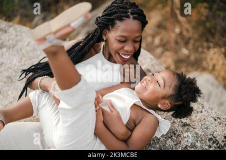 Fröhliche afroamerikanische Mutter mit lockigen Haaren umarmt und kitzelt glückliche Tochter, während Sommer Wochenende Tag auf dem Land zusammen verbringen Stockfoto