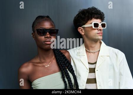 Moderne afroamerikanische Frau in trägerlosen Top und Sonnenbrille, die in der Nähe verschiedener Männer in der Nackenkette vor grauem Hintergrund steht Stockfoto
