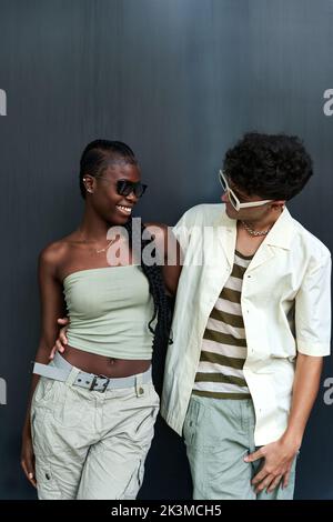 Moderne afroamerikanische Frau in trägerlosen Top und Sonnenbrille, die in der Nähe verschiedener Männer in der Nackenkette vor grauem Hintergrund steht Stockfoto
