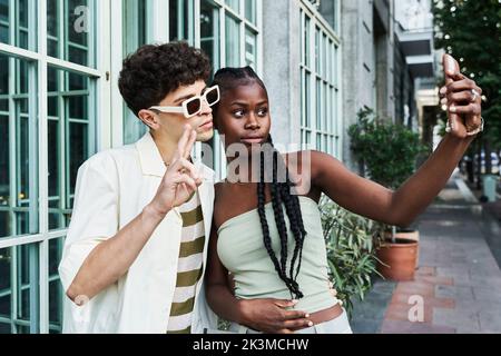 Stilvolle schwarze Frau mit langen geflochtenen Haaren, die Selfie mit einem hübschen, vielfältigen Freund in einer Sonnenbrille macht, der zwei Finger zeigt, während sie auf dem Bürgersteig steht Stockfoto