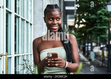 Glückliche afroamerikanische Frau mit langen Zöpfen, die eine stylische Röhrenspitze tragen und auf der Straße in der Stadt auf dem Smartphone surfen Stockfoto