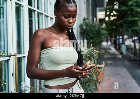 Junge afroamerikanische Frau mit langen Zöpfen, die ein stilvolles Röhrenoberteil tragen und auf der Straße in der Stadt auf dem Smartphone surfen Stockfoto