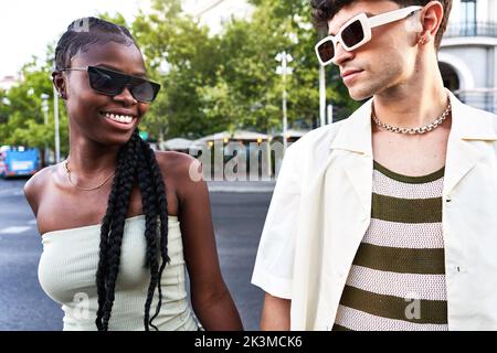 Junge, multirassische Männer und Frauen in trendigen Kleidern und Sonnenbrillen stehen auf dem Fußgängerüberweg, während sie an einem sonnigen Tag die Straße überqueren Stockfoto
