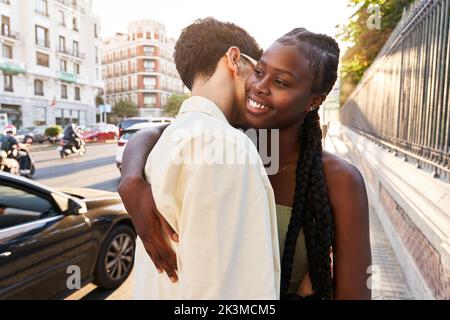 Stilvoller Mann, der eine fröhliche afroamerikanische Frau umarmt, während er während des Datums Zeit auf der Straße der Stadt verbringt Stockfoto