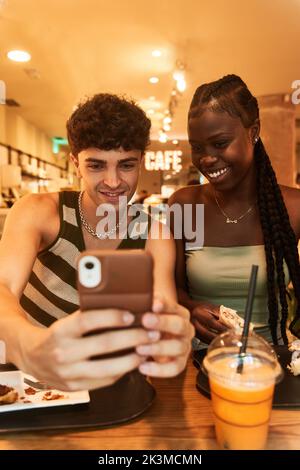 Froher junger Mann mit Smartphone in der Nähe einer schwarzen Frau, die während eines romantischen Dates im Restaurant die Rolle hält Stockfoto