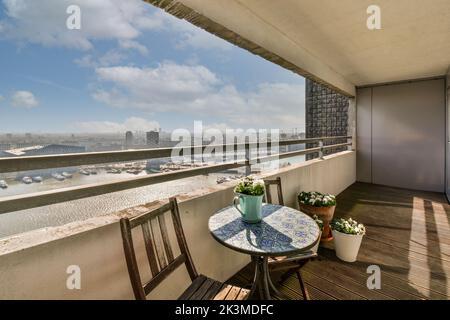 Enger langer Balkon eines modernen Apartmentgebäudes in der Nähe von Klappstühlen am Holztisch gegen ein Wohnhaus in der Stadt mit Aussicht Stockfoto