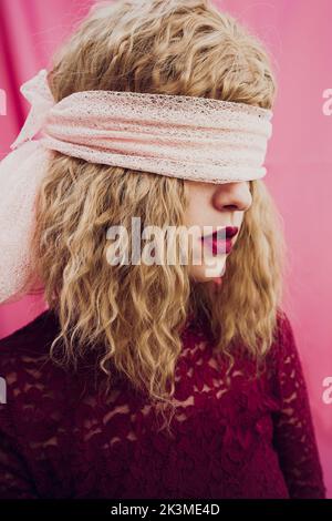 Junge Frau mit blondem Haar in Augenbinde mit roten Lippen auf rosa Hintergrund im Studio Stockfoto