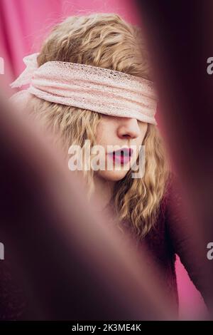 Junge Frau mit blondem Haar in Augenbinde mit roten Lippen auf rosa Hintergrund im Studio Stockfoto