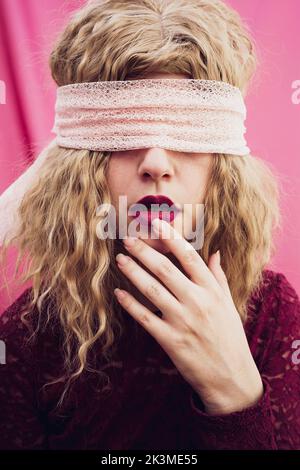 Junge Frau mit blondem Haar in Augenbinde, die Lippen mit Finger auf roten Lippen auf rosa Hintergrund im Studio berührt Stockfoto