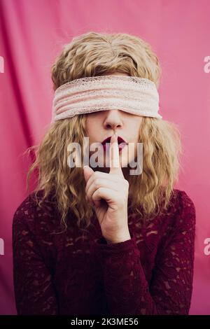 Junge Frau mit blonden Haaren in Augenbinde, die geheime Geste mit dem Finger in der Nähe roter Lippen auf rosa Hintergrund im Studio zeigt Stockfoto