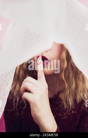 Junge, nicht erkennbare Frau, die im Studio eine geheime Geste mit dem Finger in der Nähe roter Lippen zeigt und den Kopf mit einem Schleier bedeckt Stockfoto