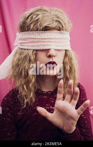 Junge Frau mit blondem Haar in Augenbinde mit roten Lippen auf rosa Hintergrund im Studio Stockfoto