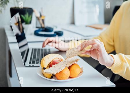 Freiberufliche junge Frau, die von zu Hause aus gesunde Lebensmittel isst. Frau, die zu Hause mit einem Laptop arbeitet und gesunde GetreideSnacks und Früchte isst Stockfoto