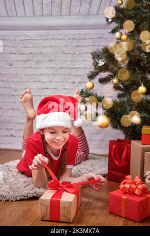 Frohe Weihnachten. Ein fröhlich lächelndes Weihnachtsmädchen in einem roten Hut und einer roten Jacke öffnet ein Weihnachtsgeschenk, liegt auf dem Boden neben dem Weihnachtsbaum. Stockfoto