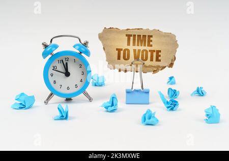 Zeitkonzept. Auf einer weißen Oberfläche, einem Wecker, einem Büroclip, zerknittertem Papier und einem Pappschild mit dem Text - Time to Vote Stockfoto