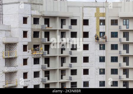 Der Prozess der Isolierung von Außenwänden in einem mehrstöckigen Gebäude im Bau. Die Arbeiter in einer orangen Konstruktionswiege befestigen die Mineralwolle s Stockfoto