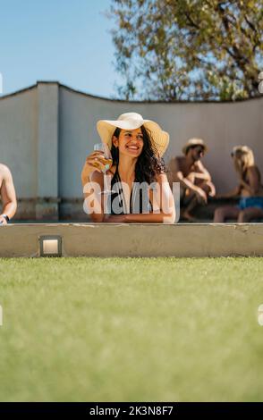 Vertikales Foto einer lächelnden jungen Frau, die eine pamela trägt und sich am Rand eines Pools lehnt Stockfoto