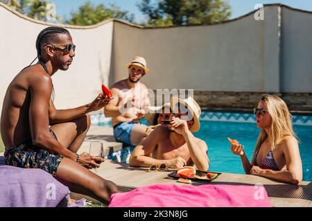 Junger schwarzer Mann mit Dreadlocks, der mit ein paar Freunden Wassermelone in der Nähe eines Schwimmbades isst Stockfoto