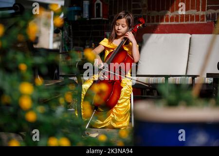 Ein kleines Mädchen in goldenem Kleid spielt im Garten draußen Cello Stockfoto