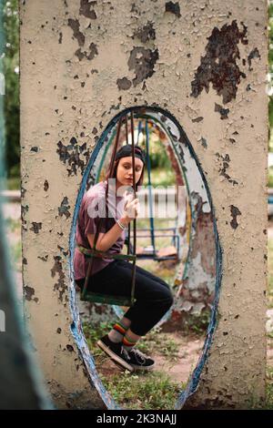 Junge Punkerin, die im Sommer auf einer Schaukel auf einem verlassenen Spielplatz sitzt Stockfoto