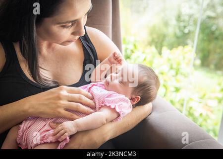 Die Frau blickt zärtlich an einem Fenster auf ihr schlafendes neugeborenes Baby in ihren Armen Stockfoto