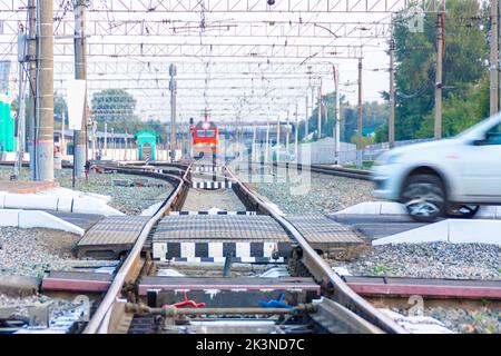 Eine rote Lokomotive der russischen Eisenbahn im Bereich eines kleinen Bahnhofs nähert sich einem Eisenbahnübergang, der gefährlich von einem Personenwagen durchquert wird, Stockfoto