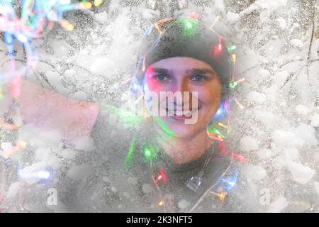 Unschärfe-Nahaufnahme-Porträt einer festlichen jungen Frau mit Girlande auf ihrem Gesicht und Kopf. Hipster Mädchen feiert weihnachten. Bokeh und Lichter. Mädchen 25 ja Stockfoto