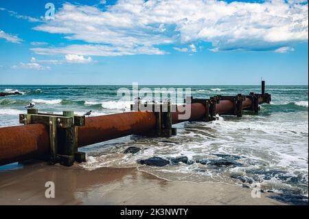 Alte rostige Wasserrohre über dem Meer mit schäumenden Wellen unter einem blau bewölkten Himmel Stockfoto