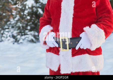 Mittlerer Ausschnitt des anonymen Weihnachtsmanns in rotem Mantel im Winterwald mit Fokus auf großen Bauch, Kopierraum Stockfoto