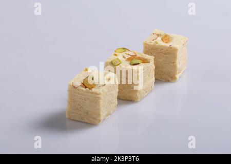 Soan Papdi ist ein beliebtes indisches würfelförmiges, knuspriges Dessert. Serviert mit Mandeln und Pistazie auf einem Teller vor launischem Hintergrund. Stockfoto