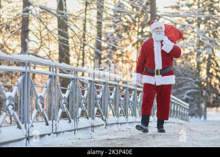 Ganzkörperporträt des traditionellen Weihnachtsmannes mit Sack und Geschenken und dem Gang zur Kamera in der Winterlandschaft, Kopierraum Stockfoto